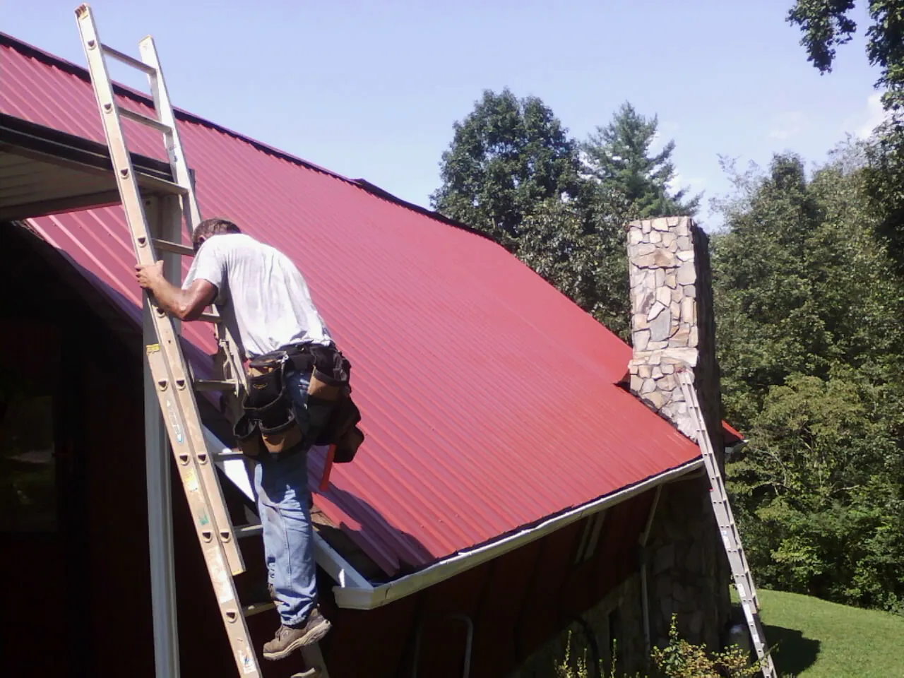 Roofer on a ladder beside a red metal roof and stone chimney — Soffit & Fascia Repair by Roofing Brothers in Falls Of Rough